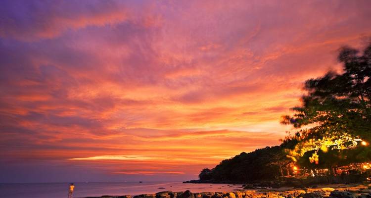 Cielo de atardecer vibrante en tonos naranjas y rosas sobre una costa tropical rocosa con árboles silueteados