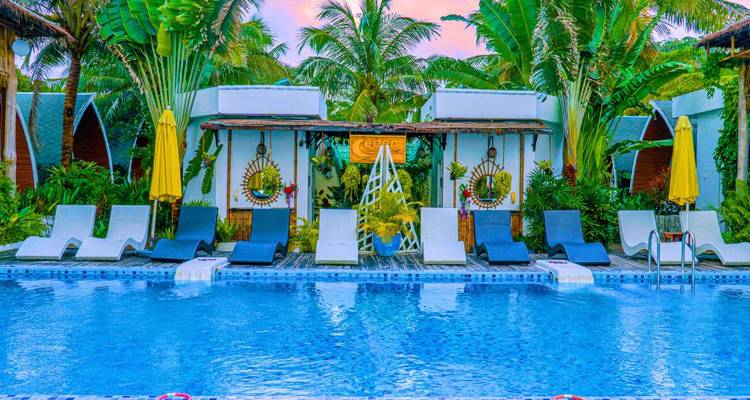 Piscina de resort tropical rodeada de palmeras, tumbonas modernas y bungalós blancos bajo un cielo rosa al atardecer.