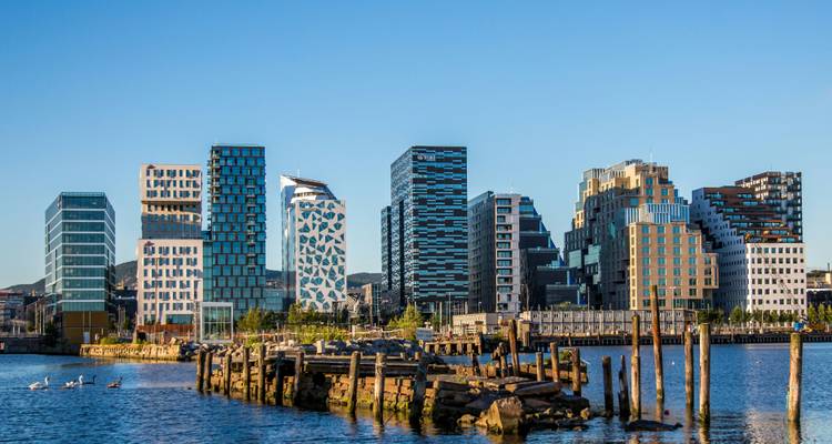Los modernos rascacielos de vidrio del barrio Barcode de Oslo elevándose detrás de un antiguo muelle de madera en una tranquila orilla fluvial.