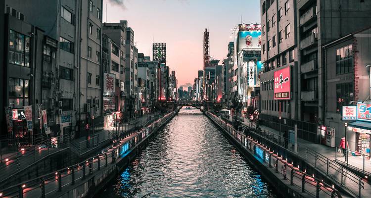 Abendliche Lichter, die sich im Wasser des Dotonbori-Kanals spiegeln, eingerahmt von Stadtgebäuden in der Dämmerung.