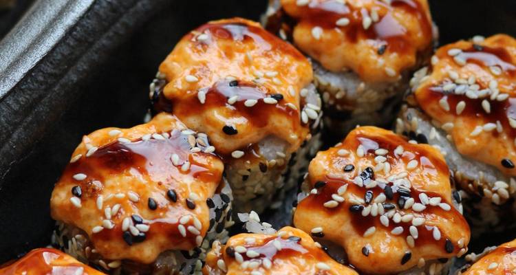 Nahaufnahme von geflammten würzigen Sushi-Rollen mit Sauce und Sesamkörnern in einer dunklen Schüssel.