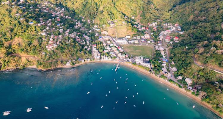 Panorama aérien d'un village côtier avec des collines verdoyantes, une baie de sable et de nombreux bateaux mouillés.