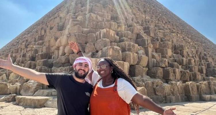 Twee reizigers met uitgestrekte armen staan voor een enorme piramide in het felle zonlicht.