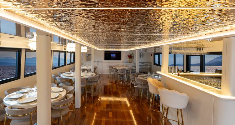 Elegante salón de comedor con pisos de madera brillante, mesas puestas, barra y grandes ventanas que enmarcan el mar.