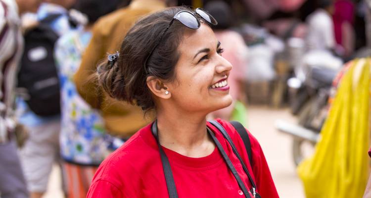 Mujer joven sonriendo con una camisa roja y gafas de sol en la cabeza parada en una calle de mercado al aire libre y concurrida.