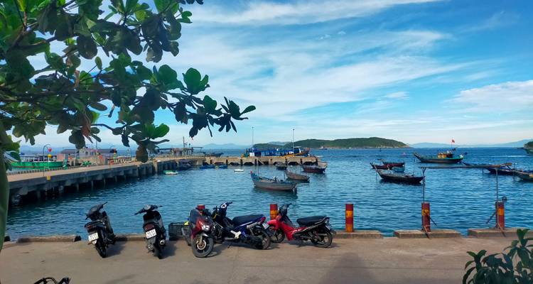 Small harbor with fishing boats, motorbikes parked on the pier and a backdrop of blue sky and islands.