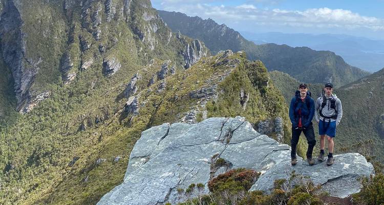 Zwei Wanderer stehen auf einer gezackten Felsenplatte und blicken auf zerklüftete Bergkämme in Tasmanien.