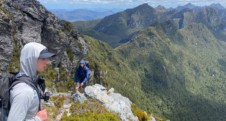 Wanderer, die ein felsiges Felsvorsprung durchqueren, mit steilen bewaldeten Bergketten im Hintergrund.