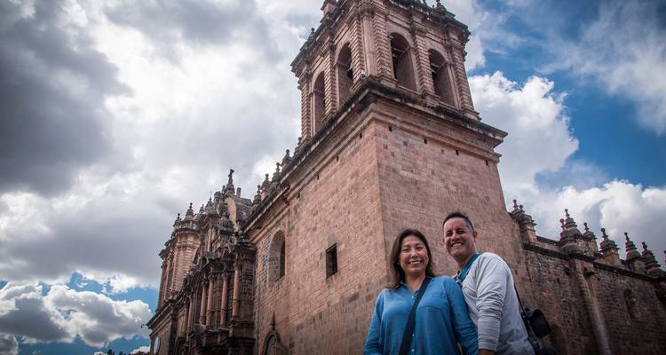Un couple souriant se tient devant une imposante tour de cathédrale en pierre sous un ciel partiellement nuageux.