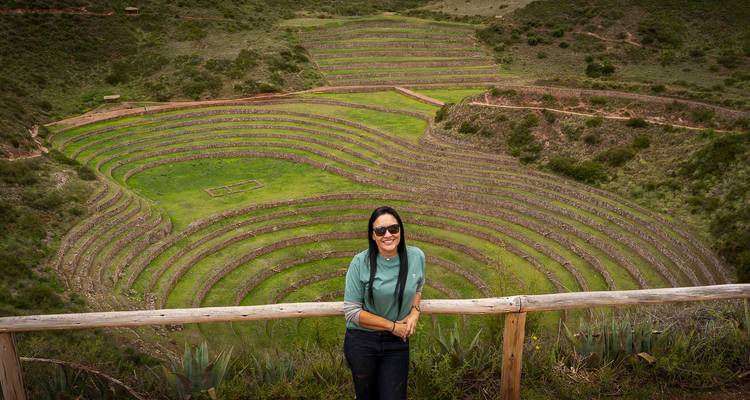 Une femme souriante s'appuie sur une rampe en bois surplombant les terrasses agricoles concentriques de Moray.