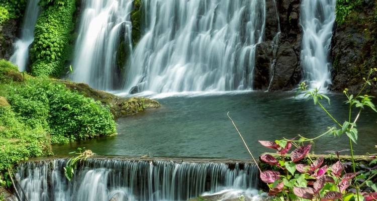 Ein mehrstufiger Wasserfall stürzt in einen klaren Pool ab, der von üppiger Vegetation umgeben ist.