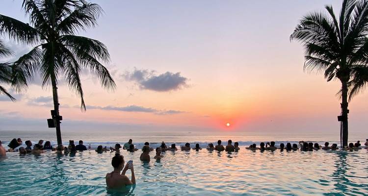 Besucherströme entspannen sich in einem Infinity-Pool zwischen Palmen, während die Sonne am Ozean-Horizont untergeht.