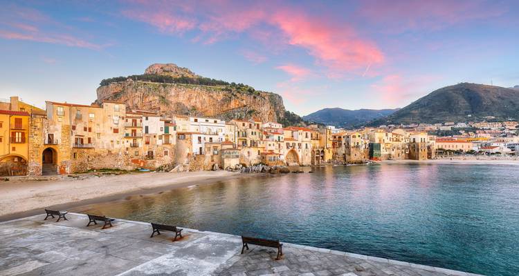 Schilderachtig kuststadje Cefalù bij roze zonsondergang dat weerspiegelt in kalm water.