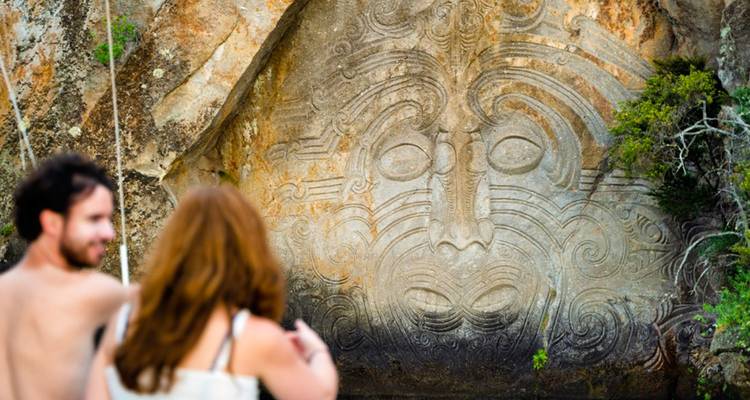 Pareja en un bote mirando una enorme escultura de roca Maorí en un acantilado a la orilla del lago