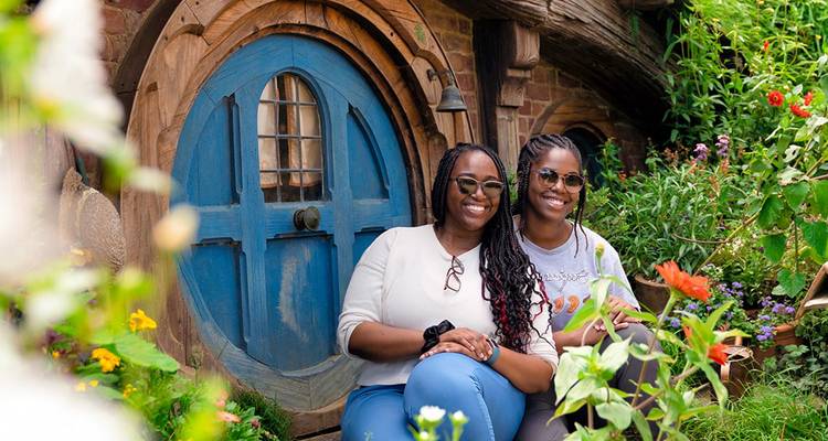 Dos visitantes sentados felizmente frente a una puerta redonda azul de hobbit rodeada de flores