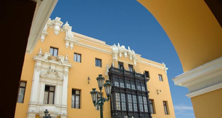 Edificio colonial amarillo enmarcado por un arco con balcón de madera ornamentada y lámparas vintage contra un cielo azul