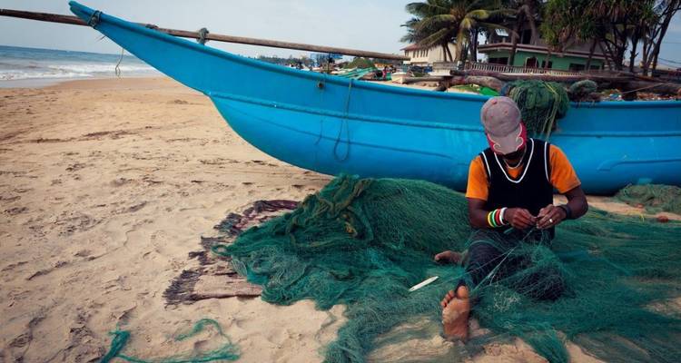 Un pescador sentado en la playa repara redes verdes al lado de una canoa con estabilizador azul con palmeras detrás.