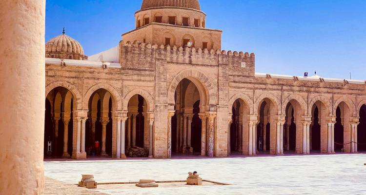 Cour de la Grande Mosquée de Kairouan avec arcades et minaret à dôme sous un ciel bleu.