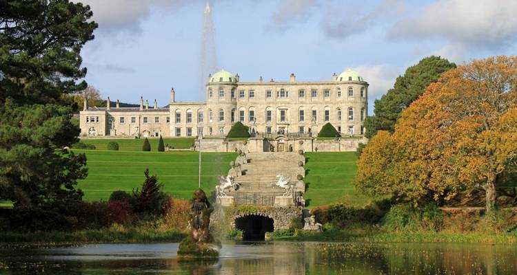 Grand Palladiaans-stijl Powerscourt House met getrapt fontijnen en beeldhouwwerk-omzoomde trappen weerspiegeld in een sieermeer te midden van herfstbomen.