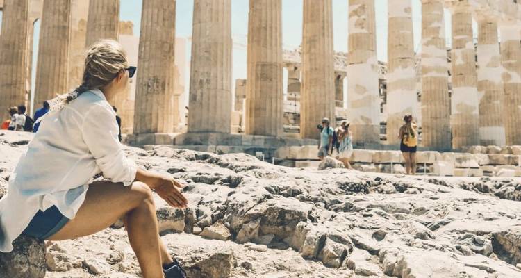 Plano general de las ruinas del Partenón con turistas y un visitante sentado en primer plano, bajo un cielo azul despejado.