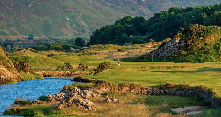 Golfeur sur un fairway verdoyant au bord d'une rivière, encadré par des collines boisées et des montagnes lointaines.
