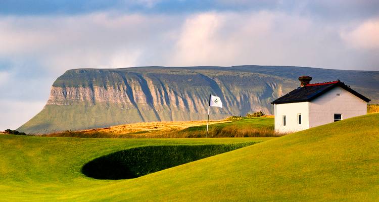Parcours de golf verdoyant avec un drapeau blanc, en arrière-plan la montagne imposante et plate de Benbulben.