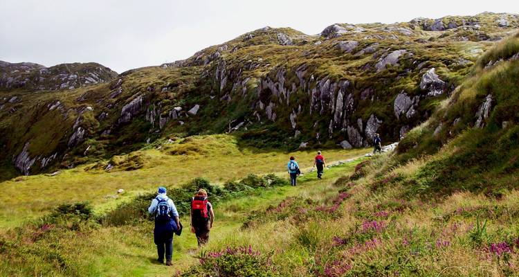 Les randonneurs avec des sacs à dos cheminent le long d'un sentier herbeux sous des collines rocheuses escarpées en Irlande.