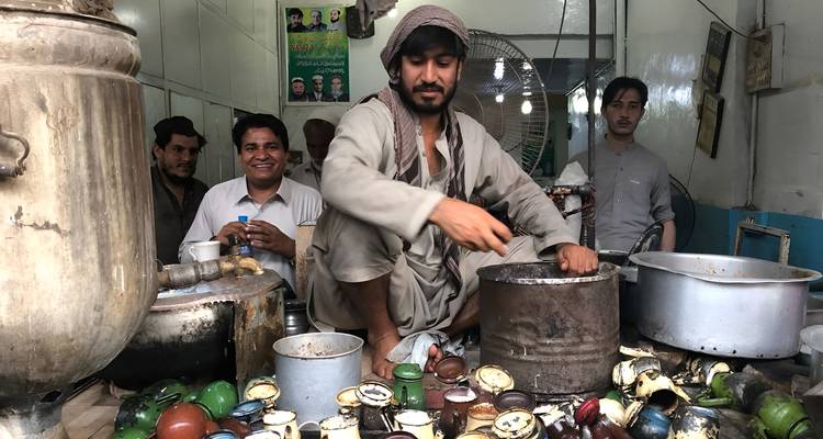 Local tea vendor squatting over steaming pots while patrons smile inside a small shop