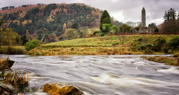 Schnell fließender Fluss vor einem bewaldeten Hügel und dem ikonischen runden Turm von Glendalough