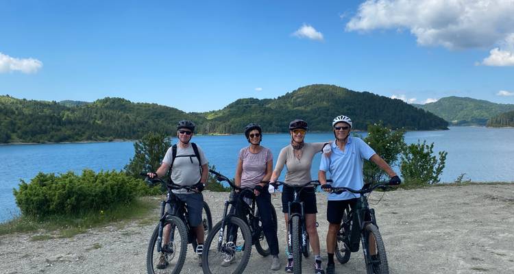 Fietsers die poseren met e-bikes naast een blauw reservoir en rollende groene heuvels onder verspreide wolken