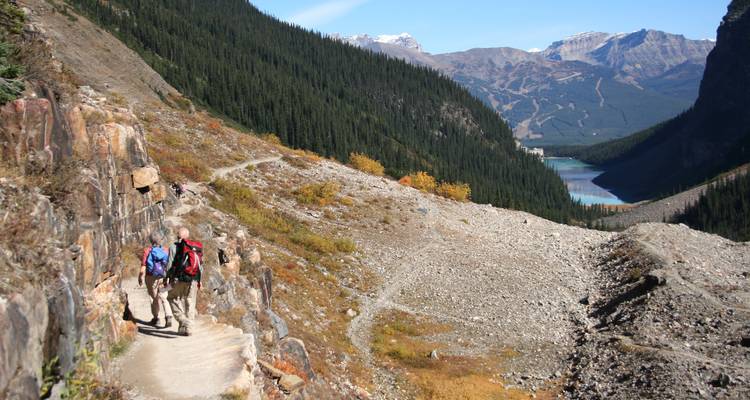 Wandelaars op een rotsachtig bergpaadje met uitzicht op een glaciaal meer en verre skipistes in het Banff National Park