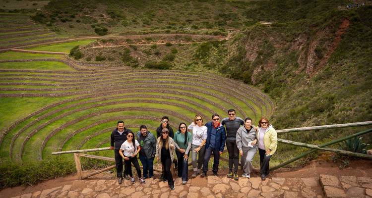 Un grupo de turistas se para en un mirador de piedra que da vista a las terrazas agrícolas concéntricas de Moray rodeadas de cerros verdes.