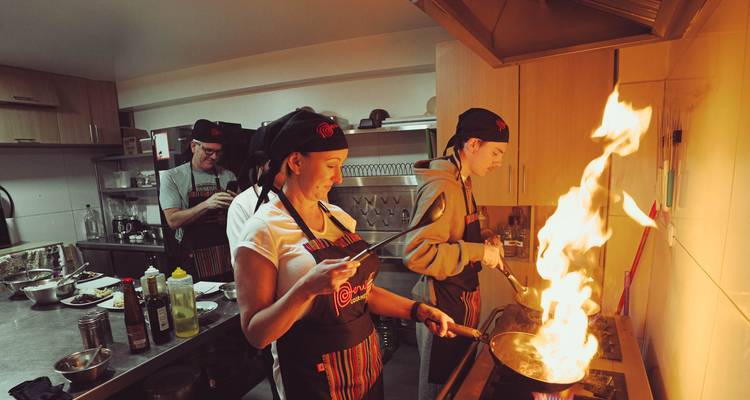 Los participantes de la clase de cocina flambean un plato en una estufa de gas en una cocina profesional, con chispas de fuego iluminando la escena.