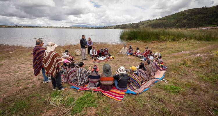 Un gran círculo de personas con ponchos se reúne junto a un lago para una ceremonia tradicional bajo un cielo nublado.