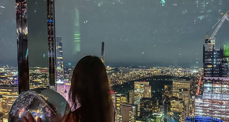 Un visiteur en silhouette contemple l'horizon nocturne de New York depuis une plateforme d'observation en verre.
