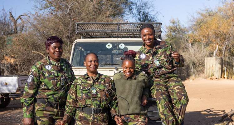 Cuatro guardaparques mujeres sonrientes en camuflaje posan frente a un vehículo de safari.