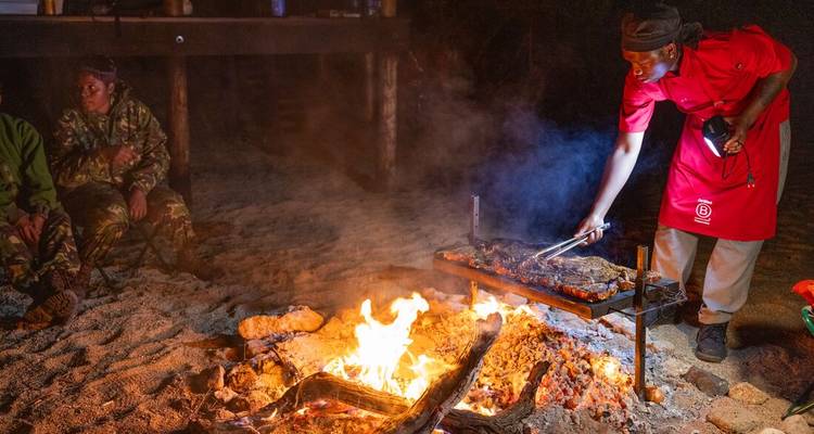 Un chef asa carne sobre una fogata abierta por la noche mientras sus compañeros se sientan cerca.
