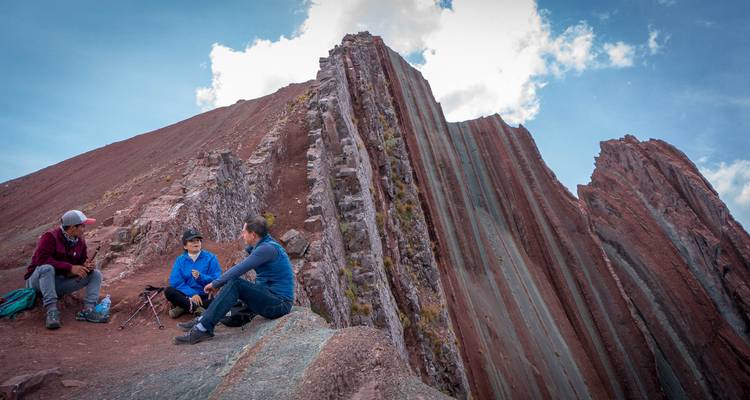 Tres excursionistas descansando en una cresta de montaña a rayas roja y verde bajo cielos brillantes en los Andes.