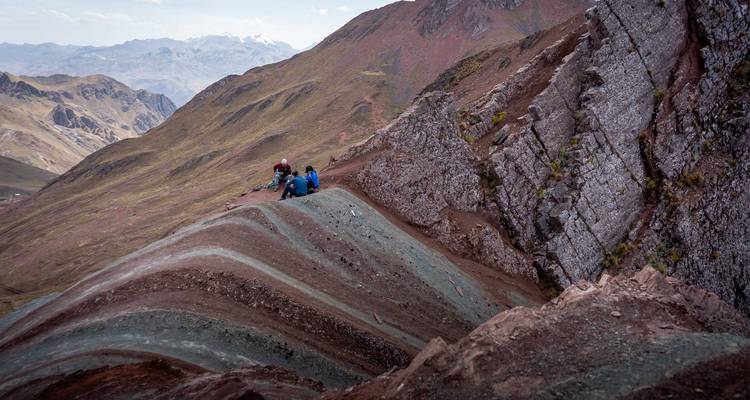 Pareja de trekkers sentados en la cima de una cresta sedimentaria multicolor con los vastos valles andinos al fondo.