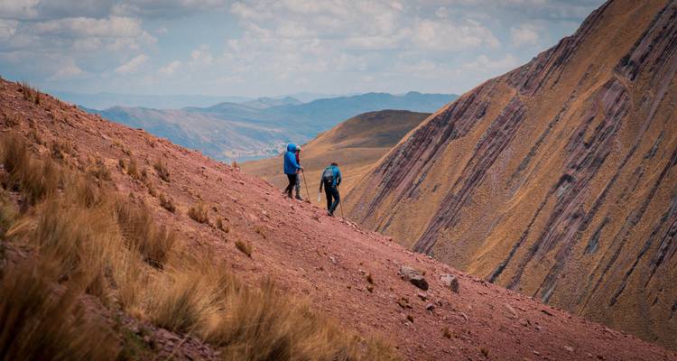 Dos excursionistas con chaquetas azules suben por una pendiente pronunciada de grava roja con acantilados rayados de fondo.