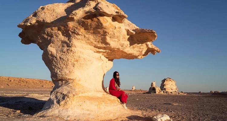 Mujer con un atuendo rojo sentada contra una gran roca blanca con forma de hongo en un desierto árido bajo un cielo azul despejado