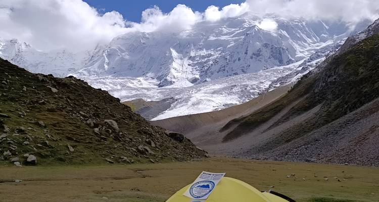 Une tente de camping jaune se dresse sur une prairie alpine sous d'imposants pics enneigés couronnés par des nuages qui dérivent.