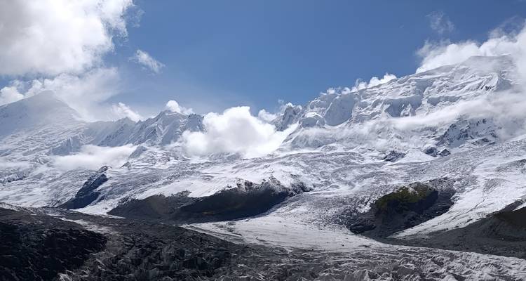 Un large glacier serpente à travers un amphithéâtre montagneux austère couvert de neige sous des nuages blanc brillant.