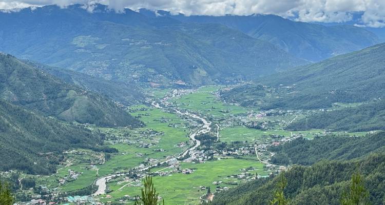 Uitgestrekt uitzicht over een vallei met een kronkelende rivier, groene rijstvelden en bergen van het Himalaya-gebergte in de verte onder de wolken.