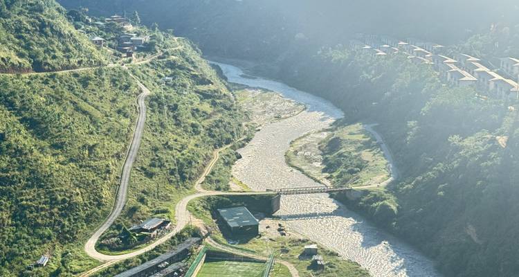 Luchtfoto van een slingerende rivier die door groene heuvels loopt, met een weg en een klein brugje in het felle zonlicht.
