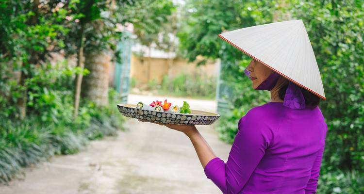 Mujer en áo dái púrpura sosteniendo una bandeja de frutas frescas en un frondoso sendero de jardín vietnamita.