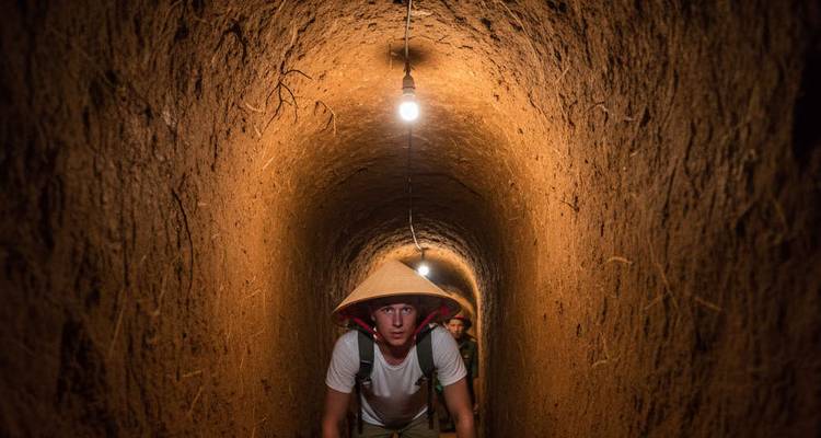 Un visitante gateando por el túnel de Cu Chi débilmente iluminado usando un sombrero cónico.