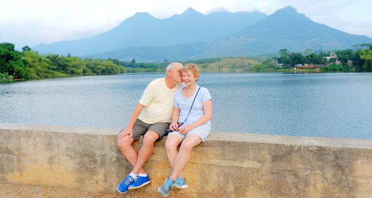 Pareja de adultos mayores sentada en una pared baja junto a un tranquilo lago con montañas azules de fondo, dándose un beso.