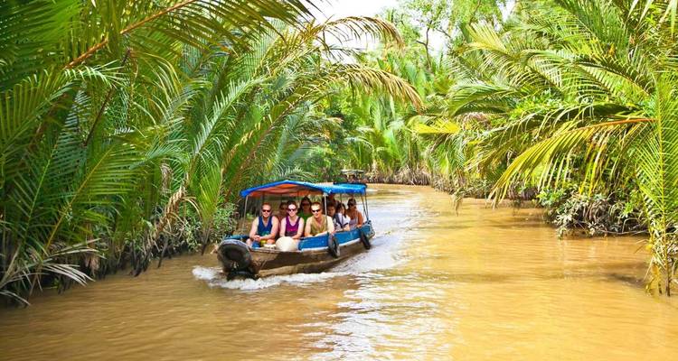 Bote de cola larga motorizado con turistas se desliza por un estrecho canal bordeado de palmeras en el Delta del Mekong.