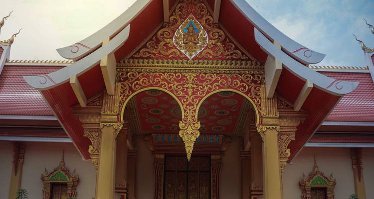 Voorgevel van een rijkelijk versierde Boeddhistisch tempel met rode en gouden details onder een zachte lucht.